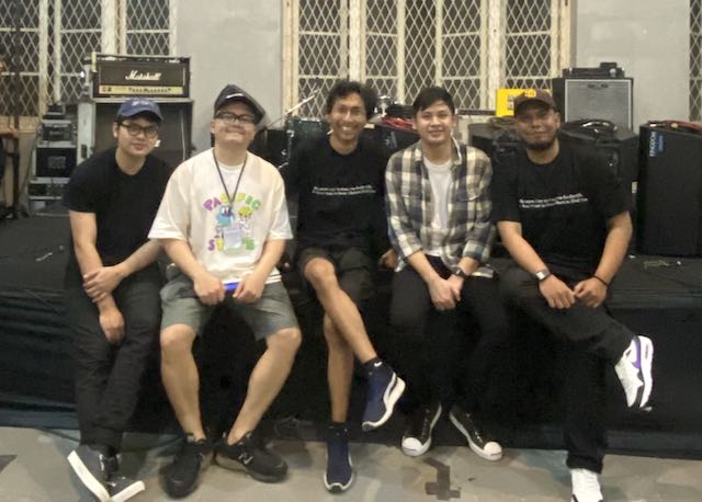 A band called eleventwelfth and Risang in the middle posing sitting in front of a small stage.